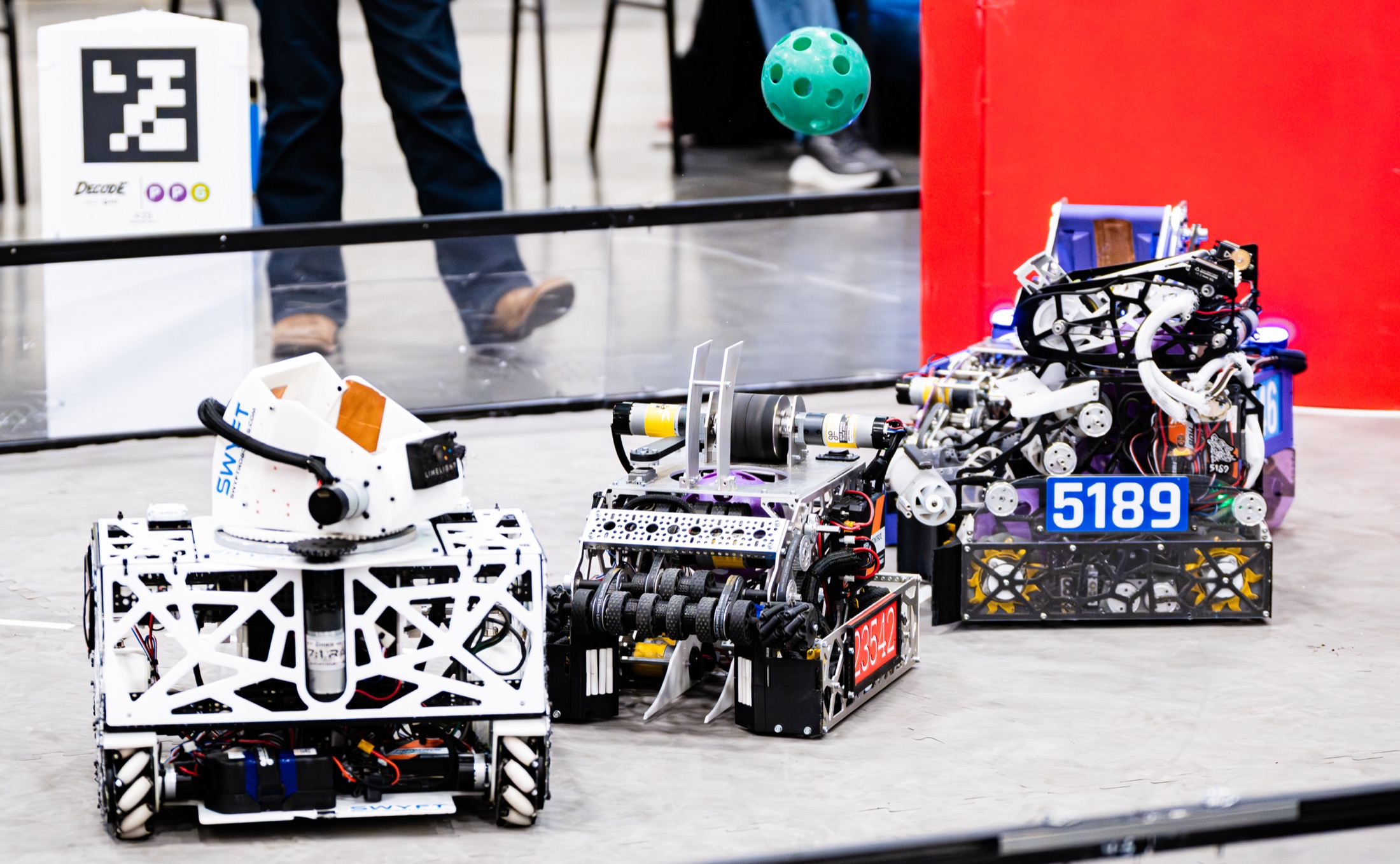 Two robots facing off on the competition field mid-match, an AprilTag and a green game ball in frame