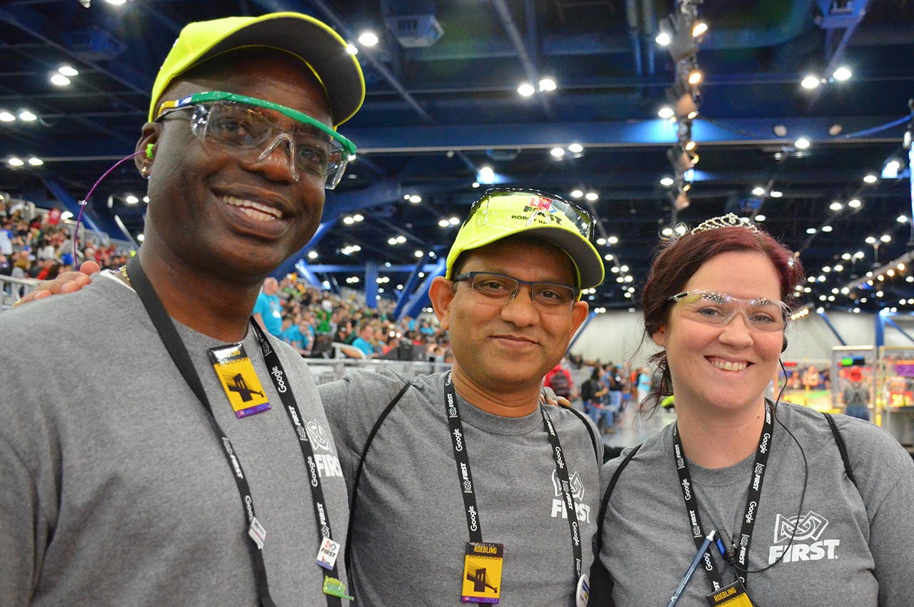 Three event volunteers in FIRST shirts and caps at a competition venue