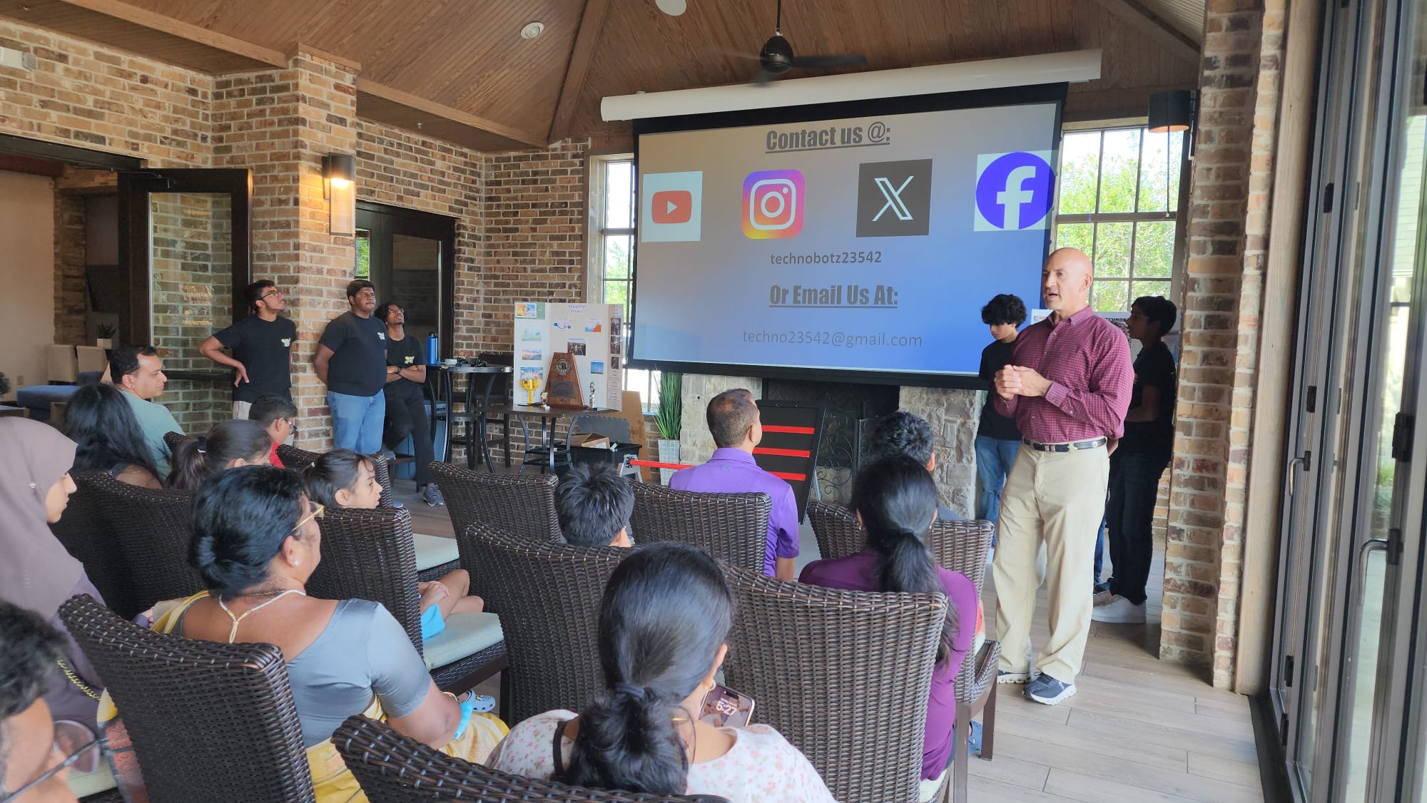 Texas State Representative Matt Shaheen speaking to a seated audience at a TechnoBotz outreach event, the team's contact details on the screen behind him