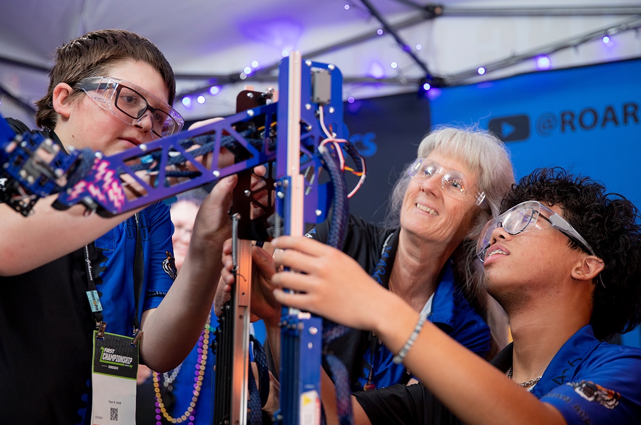 A mentor and two students in safety glasses working together on a tall robot mechanism