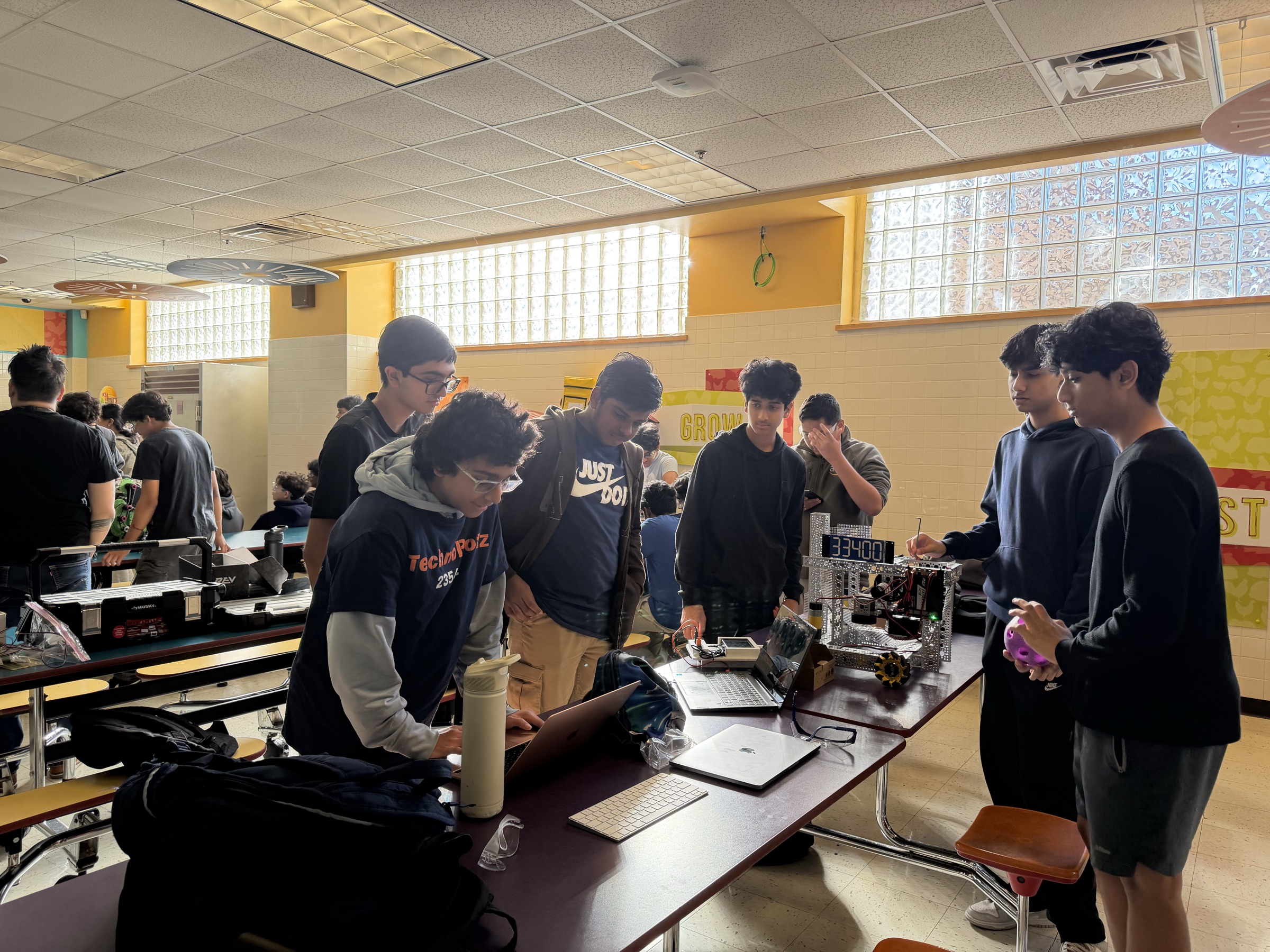 Students from several teams around cafeteria tables at a build session, laptops open and robots on the bench