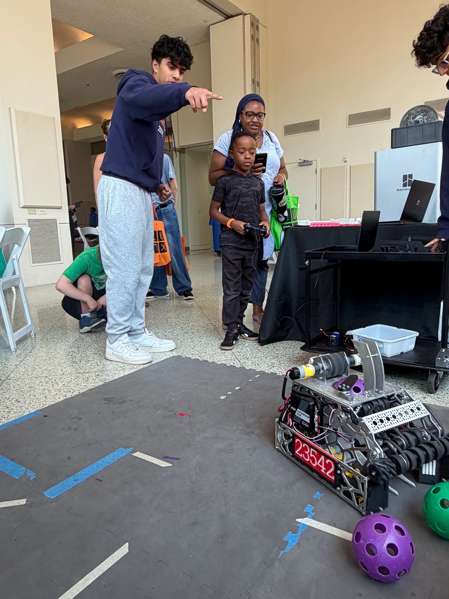 A TechnoBotz student coaching a young child driving the competition robot at an outreach event while a parent films