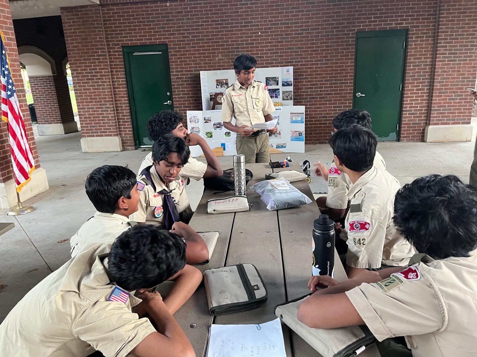 A TechnoBotz student presenting a display board to scouts seated around a picnic table, robot parts laid out in front of them