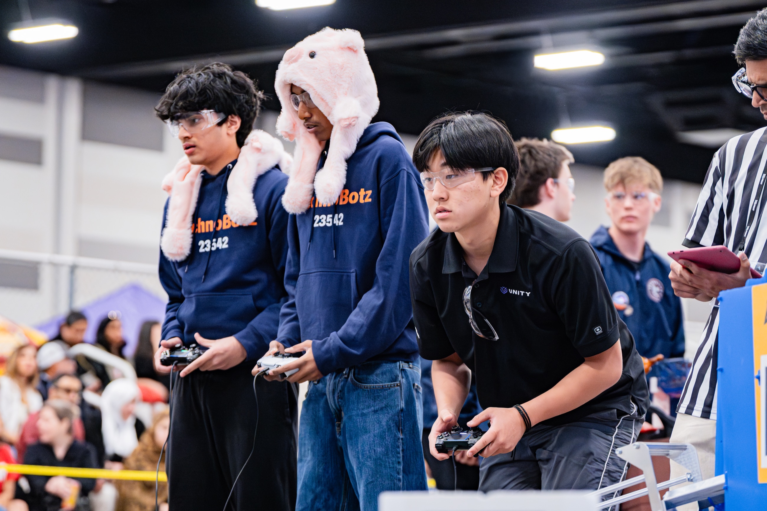 Three TechnoBotz drivers at the field holding controllers, two in team hoodies and one in a pink pig hat