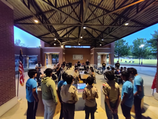 Students in TechnoBotz shirts running a robot demonstration for a scout troop under a pavilion at dusk